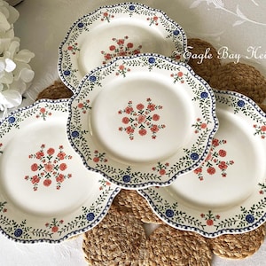 May include: Four white ceramic plates with scalloped edges, adorned with a floral pattern in red and blue. The plates feature a blue border with a floral and leaf design. The text "Eagle Bay Heirlooms" is visible.