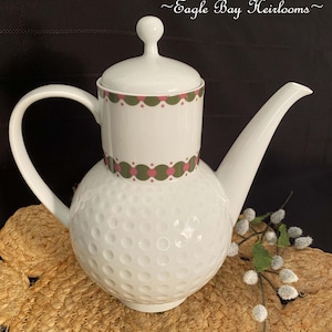 May include: White ceramic teapot with a golf ball texture on the bottom half and a cylindrical top with a handle and lid. The top has a decorative band of pink and green circles. The text "~Eagle Bay Heirlooms~" is in the upper right corner.