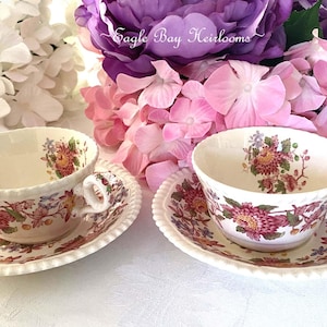 May include: Two cream-colored ceramic teacups and matching saucers featuring a detailed floral pattern in shades of pink, red, and green. Each piece has a scalloped, beaded edge and a decorative handle. The text Eagle Bay Heirlooms is visible.