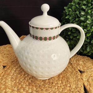 May include: White ceramic teapot with a golf ball-like texture and a lid with a decorative band of pink and green circles. The teapot has a curved spout and handle. The teapot sits on a woven coaster.