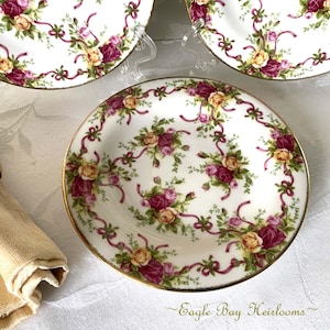 May include: White porcelain plates with a floral rose pattern in shades of pink, yellow, and green, accented with burgundy ribbons and gold trim. The plates are part of a set, perfect for serving desserts or appetizers. The text "Eagle Bay Heirlooms" is at the bottom.