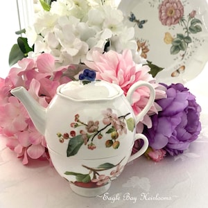 May include: A white ceramic tea-for-one set featuring a teapot stacked on a teacup, decorated with a botanical print of pink apple blossoms, green leaves, and small fruits. The teapot lid has a small blue fruit-shaped handle.