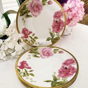 May include: Set of four white porcelain plates with a gold rim and a floral rose design. The plates feature pink and red roses with green leaves. The text "Eagle Bay Heirlooms" is at the bottom.