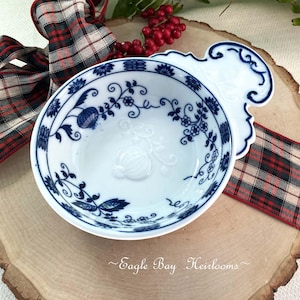May include: White porcelain bowl with blue floral design and a decorative handle. The bowl features a detailed pattern of flowers and leaves. The rim and handle are outlined in blue. The bowl is on a wooden surface with a plaid ribbon.