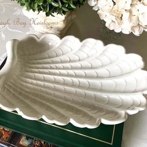 May include: A white, shell-shaped ceramic serving dish with a scalloped edge. The dish has a textured, ribbed design and sits on a green book. The words "~Eagle Bay Heirlooms" are visible in the background.