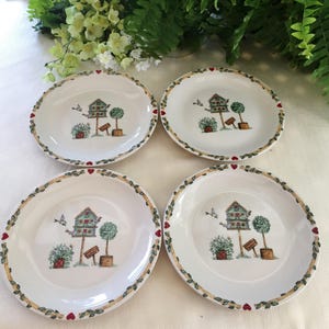May include: Set of four white ceramic salad plates with a green and yellow border. Each plate features a birdhouse with a red roof and two small trees. The plates have a red heart design on the border.