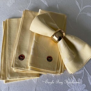 May include: A set of pale yellow linen napkins with a textured pattern, accented with a gold napkin ring. Each napkin has a small, round emblem. The napkins are neatly stacked, suggesting formal dining.