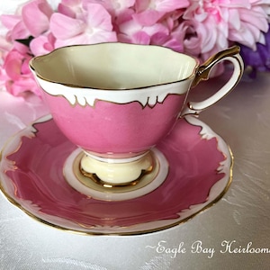 May include: A pink teacup and saucer set with gold trim and a white scalloped edge. The teacup has a matching handle and sits on a cream-colored base. The background features pink and purple flowers.