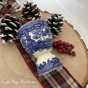 May include: A blue and white ceramic egg cup with a Willow pattern design. The egg cup is displayed on a wooden slice with pine cones and a red and green plaid ribbon. The text "Eagle Bay Heirlooms" is at the bottom.