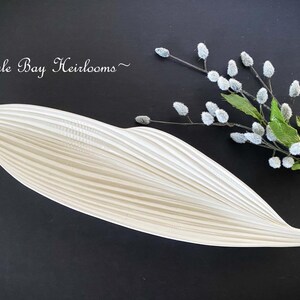 May include: A cream-colored, leaf-shaped decorative dish with a ribbed texture. The dish is positioned on a dark surface, accompanied by a sprig of faux flowers with white buds and green leaves. The text "~Eagle Bay Heirlooms~" is in the upper left corner.