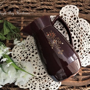 May include: A dark brown ceramic pitcher with a floral design in gold and orange. The pitcher has a wide mouth and a handle.