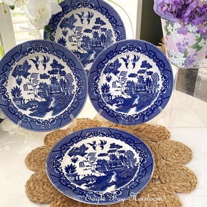 May include: Four blue and white Willow pattern plates on a table. The plates have a detailed landscape design with a blue border. The plates are displayed on a woven placemat and clear stands. The plates are surrounded by flowers.
