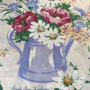 May include: Floral print fabric featuring a blue watering can filled with a bouquet of flowers. The flowers include white daisies, red poppies, and blue cornflowers. The text "Eagle Bay Heirlooms" is printed at the bottom.