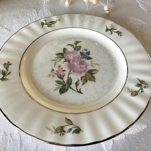 May include: A white porcelain plate with a gold rim and a floral design. The center of the plate features a bouquet of pink roses and green leaves. The plate has a fluted edge and small floral accents.