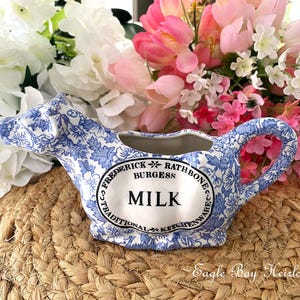 May include: A blue and white ceramic milk pitcher with a floral design and the word "MILK" in the center. The pitcher is surrounded by pink and white flowers, and sits on a woven surface.