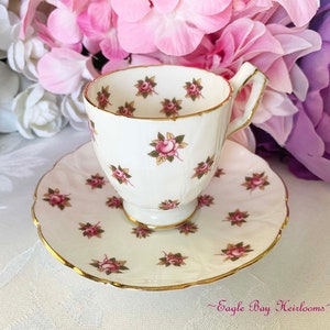 May include: A white teacup and saucer with a delicate floral pattern of pink roses and green leaves. The cup and saucer have a gold trim.