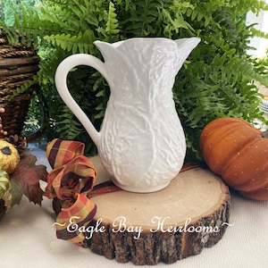 May include: A white ceramic pitcher featuring a textured cabbage leaf pattern on its exterior surface. The pitcher has a curved handle and a scalloped rim, with the text Eagle Bay Heirlooms written in white script across the front base area.