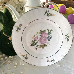 May include: A white porcelain plate with a floral design featuring pink and purple roses. The plate has a silver rim and is displayed on a clear stand. Easter eggs in a basket and a green bunny are in the background.
