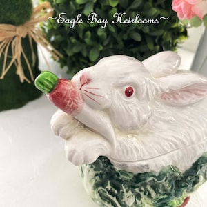May include: A white ceramic bunny-shaped container with a carrot in its mouth. The bunny has red eyes and pink accents on its ears and nose. The base is green and white. The text "Eagle Bay Heirlooms" is visible.