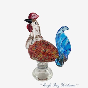 May include: A clear glass rooster figurine with a red comb and wattle, a black beak, and a blue tail. The body is a speckled mix of red, yellow, and brown. The figurine sits on a clear glass base. The text "Eagle Bay Heirlooms" is at the bottom.
