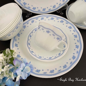 May include: A white porcelain dinnerware set with a blue floral pattern. The set includes plates, saucers, bowls, and teacups. The edges of the plates and bowls have a scalloped design. The text "Eagle Bay Heirlooms" is visible.