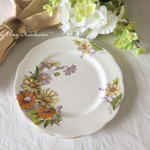 May include: A white ceramic plate with a scalloped edge features a floral design of yellow, orange, and purple flowers with green leaves. The plate has a gold rim.