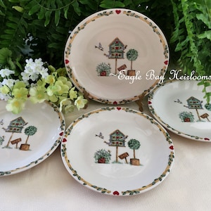 May include: Set of four white ceramic plates with green and brown floral designs. Each plate features a birdhouse with a red roof and a small tree. The plates have a red heart border.