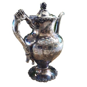 May include: A silver teapot with an ornate design featuring grapes and vines. The teapot has a lid with a finial in the shape of a bunch of grapes. The teapot has a round body and a flared base.