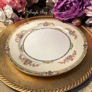 May include: A white porcelain dinner plate with a floral pattern in shades of pink, blue, and green. The plate has a gold rim and is sitting on a gold charger plate.