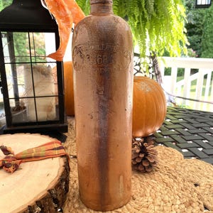 May include: A tall, brown antique bottle with embossed text, likely stoneware. The bottle has a cylindrical shape and a narrow neck. The background includes pumpkins, a lantern, and decorative accents, suggesting a fall theme.