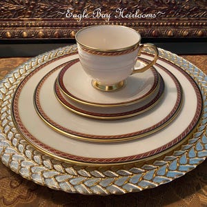 May include: A set of white porcelain dinnerware with gold and burgundy trim. The set includes a cup, saucer, and two plates. The plates have a woven pattern.