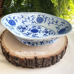 May include: A white ceramic serving dish with a blue floral design. The dish has a scalloped edge and a raised base. It is sitting on a wooden slice.