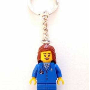 以下が含まれることがあります： 茶色の髪、黄色の顔、小さな飛行機のロゴが付いた青い制服を着た、青いレゴミニフィギュアの客室乗務員。ミニフィギュアは銀色のキーホルダーに取り付けられています。