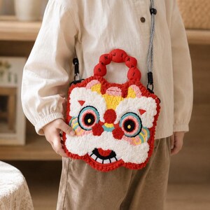 May include: A red, white, and yellow crossbody bag with a lion dance design. The bag features a detailed, embroidered lion face with large eyes, a smiling mouth, and a red handle. The strap is black and white.