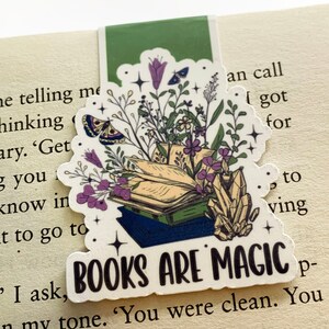 Könnte beinhalten: Ein weißer Aufkleber mit einem Stapel Bücher, Blumen, Schmetterlingen und Kristallen. Der Aufkleber trägt den Text "BOOKS ARE MAGIC" in schwarzen Buchstaben.