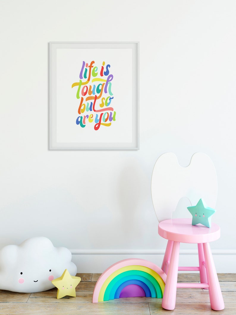 May include: A framed print with the text "life is tough but so are you" in colorful lettering. The print is hanging on a white wall above a pink wooden stool with a white cloud-shaped toy and a rainbow-colored wooden toy.