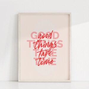 May include: Framed art print with the words "GOOD THINGS TAKE TIME" in red and pink lettering. The artwork is displayed in a light-colored wooden frame against a white background. The overall aesthetic is minimalist and inspirational.