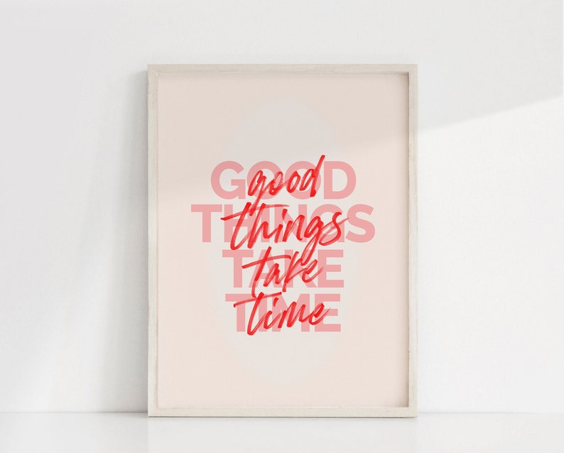 May include: Framed art print with the words "GOOD THINGS TAKE TIME" in red and pink lettering. The artwork is displayed in a light-colored wooden frame against a white background. The overall aesthetic is minimalist and inspirational.