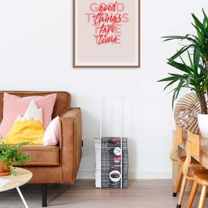 May include: A framed print with the words "Good Things Take Time" in red and pink lettering hangs above a brown leather sofa with colorful pillows. A stack of magazines is next to the sofa. A potted plant and wooden table are also in the room.