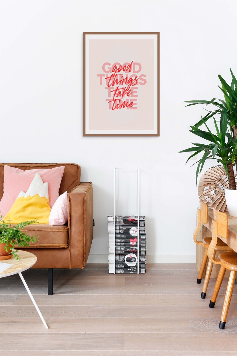 May include: A framed print with the words "Good Things Take Time" in red and pink lettering hangs above a brown leather sofa with colorful pillows. A stack of magazines is next to the sofa. A potted plant and wooden table are also in the room.