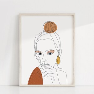 以下が含まれることがあります： 髪をまとめた女性の線画。女性は茶色と黄色の服を着て、黄色のイヤリングをしています。絵は白い額縁に入っています。