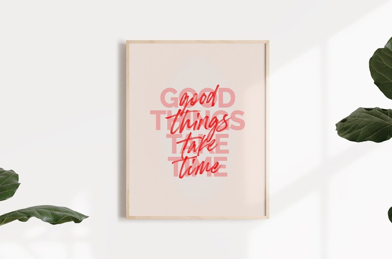 May include: A framed print with the words "GOOD THINGS TAKE TIME" in red and pink lettering. The print has a light beige background and a natural wood frame. The image is set against a white wall with a plant in the corner.