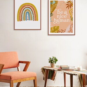 May include: Two framed prints on a white wall. The print on the left features a rainbow with a textured design in shades of brown, yellow, blue, and white. The print on the right features a peach background with a leafy design and the text "be a nice human".