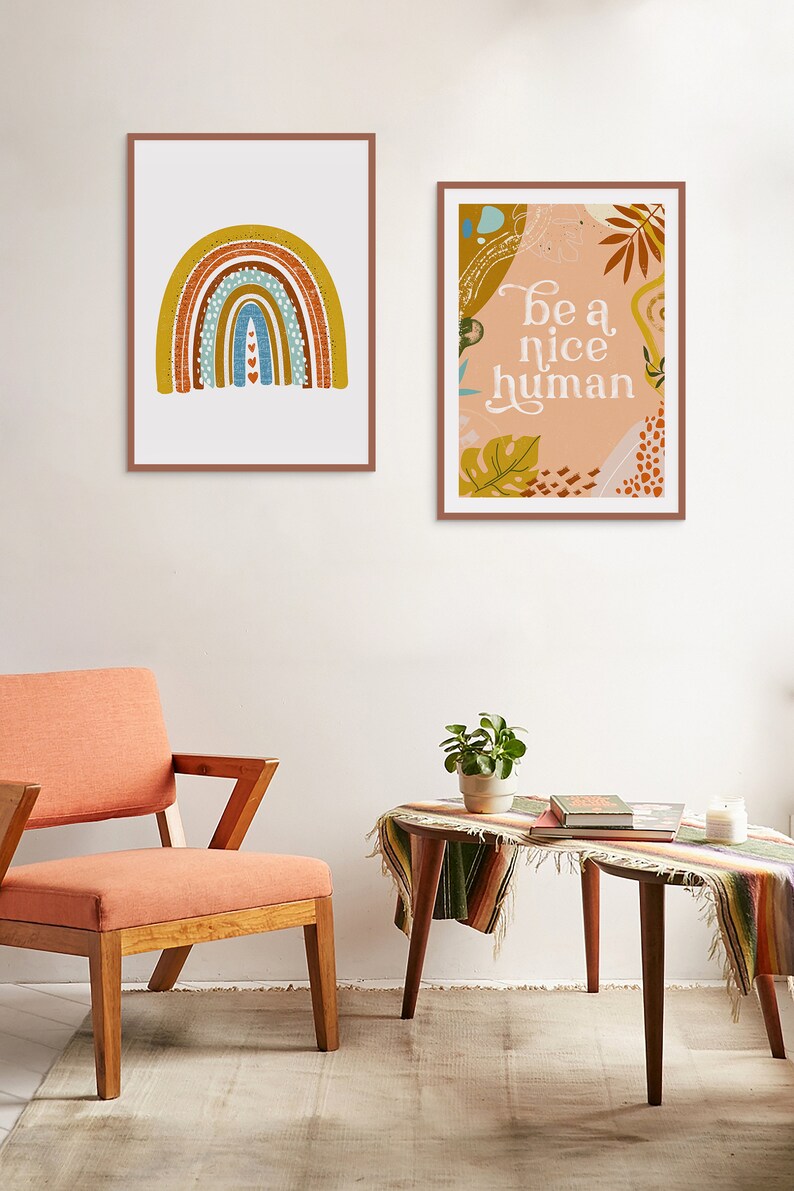 May include: Two framed prints on a white wall. The print on the left features a rainbow with a textured design in shades of brown, yellow, blue, and white. The print on the right features a peach background with a leafy design and the text "be a nice human".