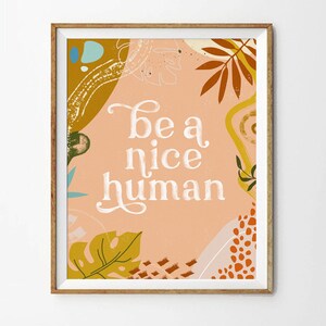 May include: A peach-colored poster with a white background and the text "be a nice human" in a white cursive font. The poster is decorated with abstract shapes and leaves in shades of orange, yellow, and green.