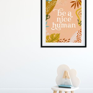 May include: A framed print with a peach background and abstract shapes in shades of brown, yellow, and green. The text "be a nice human" is written in white cursive font.