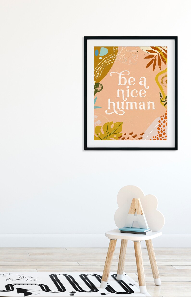 May include: A framed print with a peach background and abstract shapes in shades of brown, yellow, and green. The text "be a nice human" is written in white cursive font.