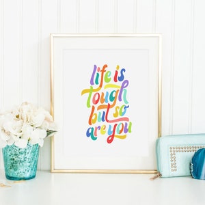 May include: A gold framed print with the text "life is tough but so are you" in colorful lettering on a white background. The print is sitting on a white surface with a white hydrangea in a blue vase and a light blue clutch purse with a blue agate stone.