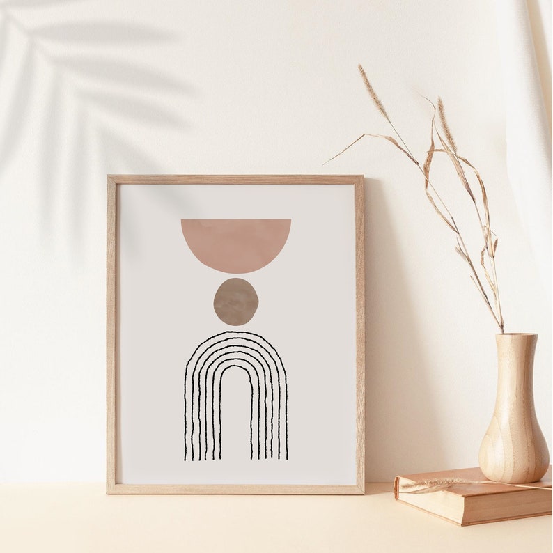 May include: A framed print with a minimalist abstract design featuring a pink half-circle, a brown circle, and a black arch with vertical lines. The print is on a white background and is framed in a light wood frame.