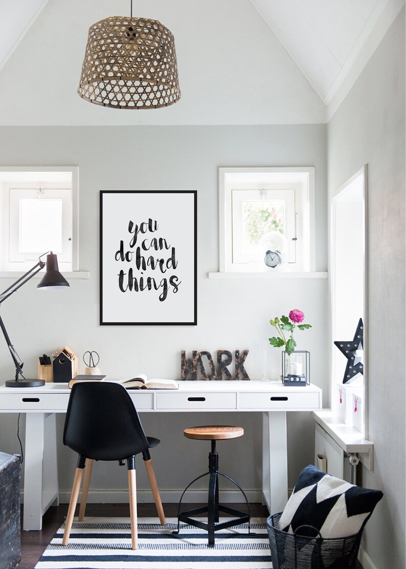 Op de afbeelding: Een wit bureau met drie lades en een zwarte stoel. Het bureau heeft een motiverende quote aan de muur erboven die zegt "you can do hard things". Er staat een houten kruk naast de stoel en een zwart-wit gepatroneerd kussen in een zwarte mand op de vloer.